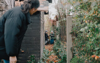 Mutter spielt Verstecken mit ihren Söhnen, die Mutter entdeckt sie hinter der Gartenhütte Natürliche dokumentarische Familienfotografie - Maria Lea Photography